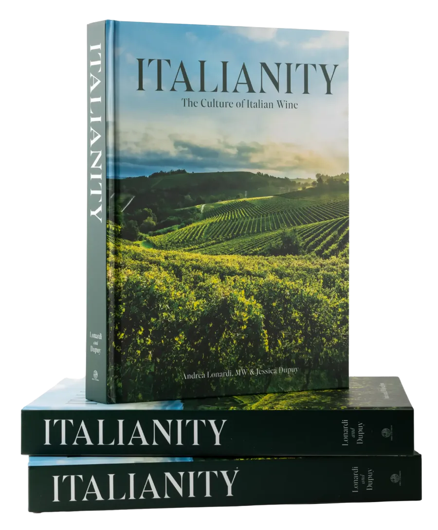 The hardcover Italianity: The Culture of Italian Wine standing upright on two copies lying flat, its cover showing vineyards under a bright sky.